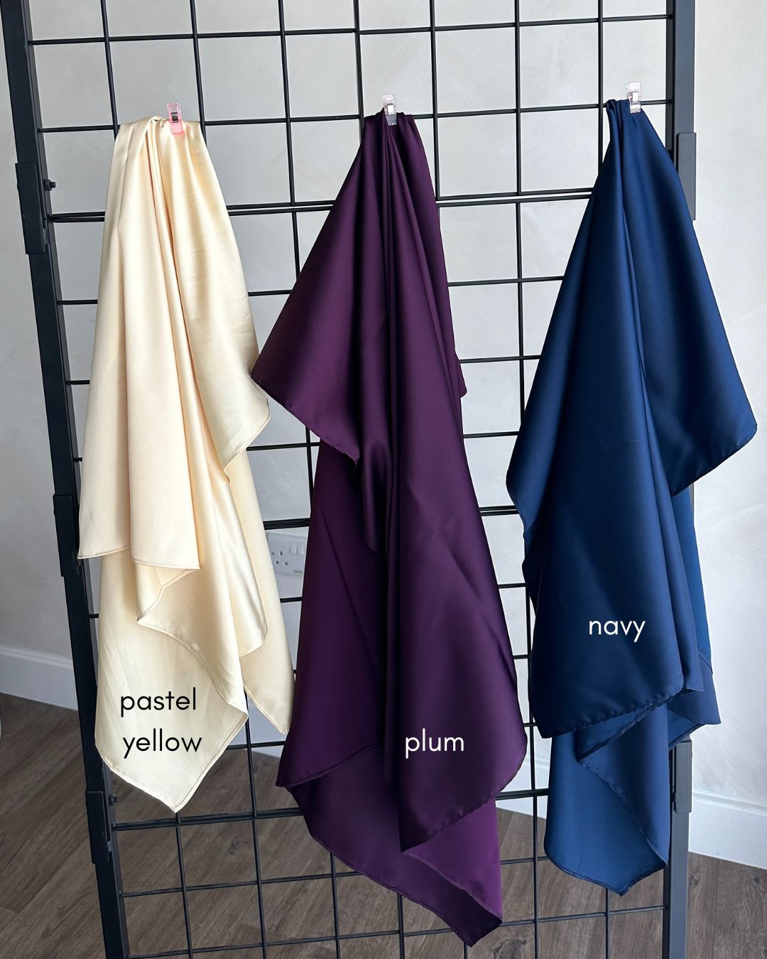 Three scarves in butter yellow, plum, and navy hanging on a rack against a tiled wall.